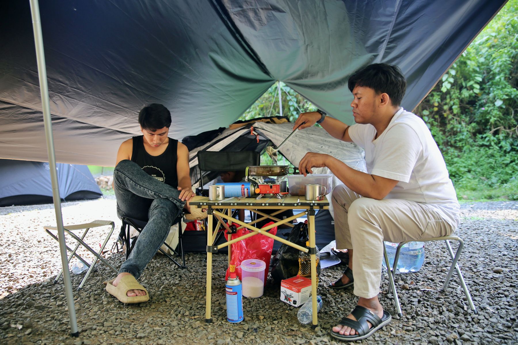 Two campers cooking at a folding table under a tarp shelter beside their tent