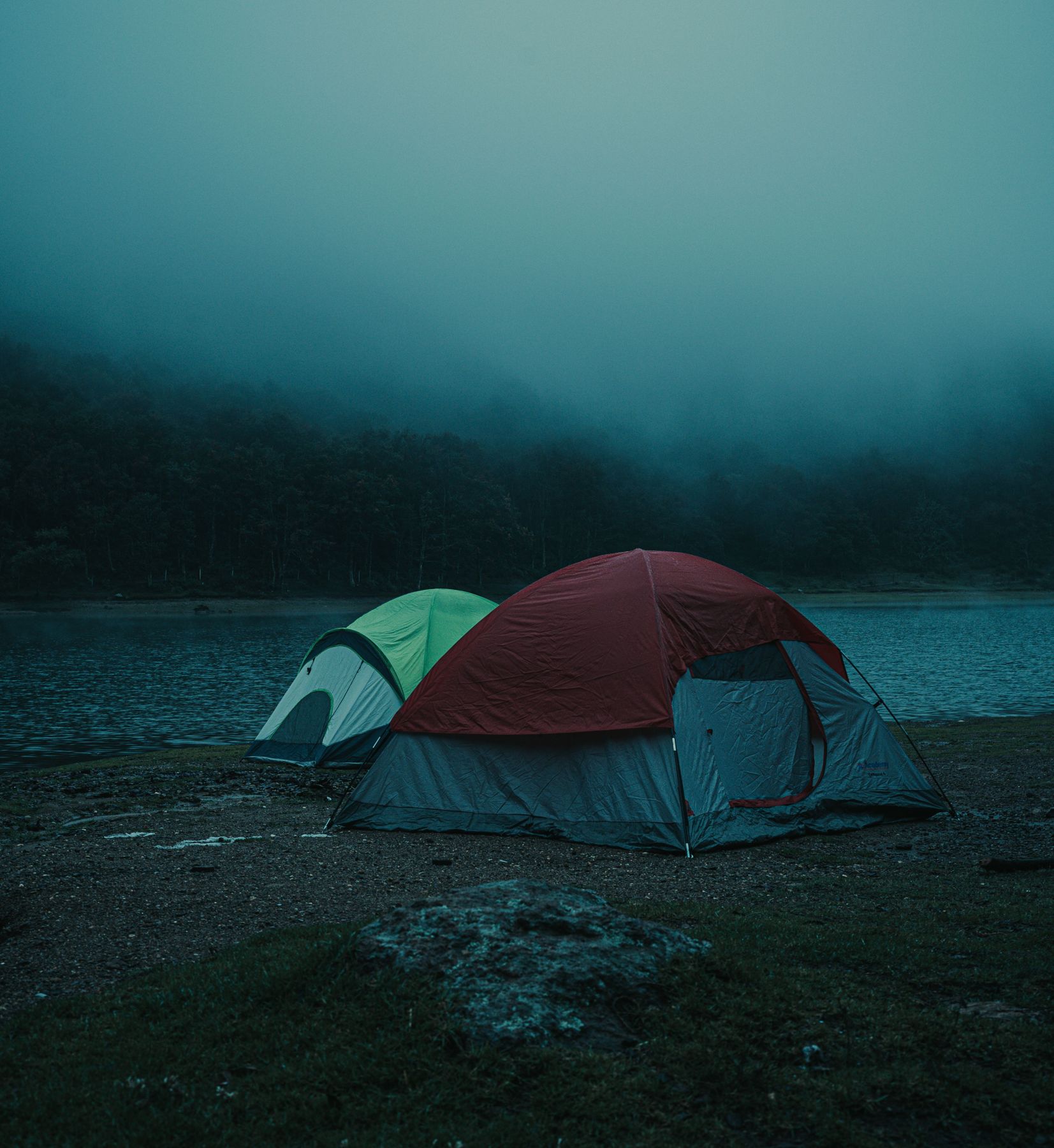 Red and green dome tents on a misty lakeshore with forested hills behind