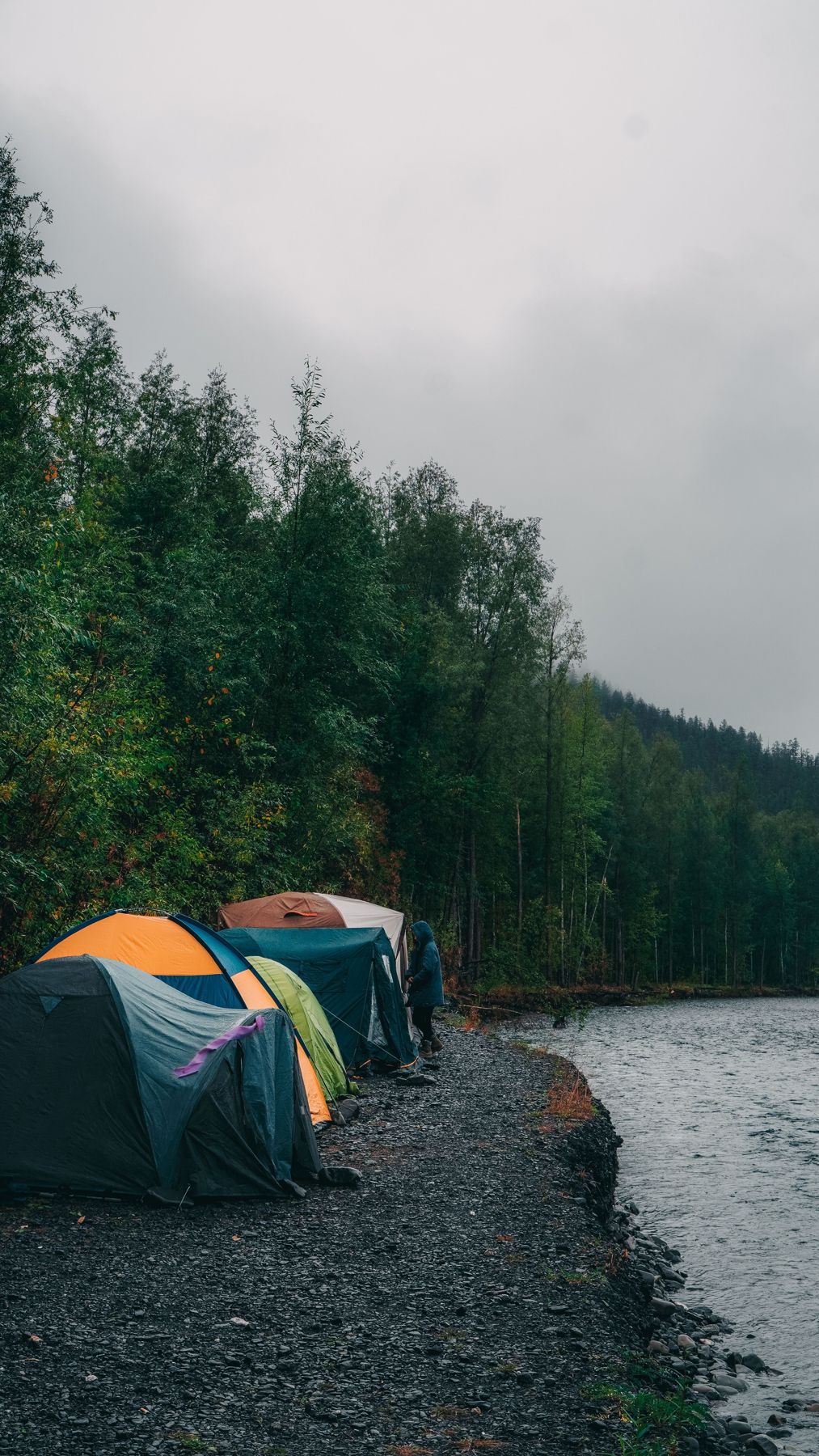 Row of colourful tents along a gravel riverbank under grey rainy skies